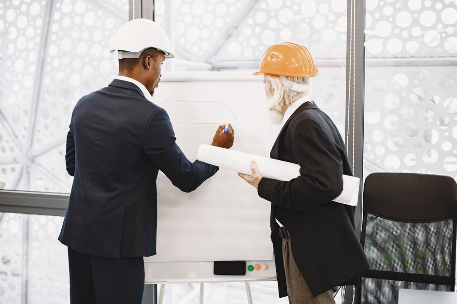 Dois arquitetos de capacete trabalhando em uma planta, destacando trabalho em equipe
